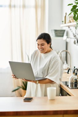 Genç bir kadın evdeki geniş, aydınlık bir mutfakta laptopunu kullanırken gülümsüyor..
