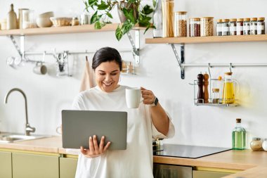 Bright kitchen with a cheerful young plus sized woman sipping coffee and using her laptop. clipart