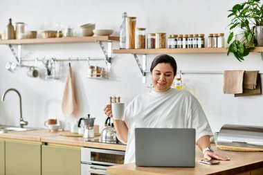 Sıcak bir mutfakta, iri yarı genç bir kadın elinde kupa tutarken ve laptopunu kullanırken gülümsüyor.