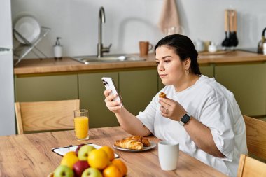Genç ve iri yarı bir kadın mutfakta akıllı telefonuyla uğraşırken kahvaltının tadını çıkarıyor..