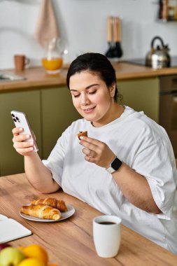 Genç, iri yarı bir kadın, telefonuna gülümseyen, parlak, modern bir mutfakta hamur işlerinin tadını çıkarıyor..