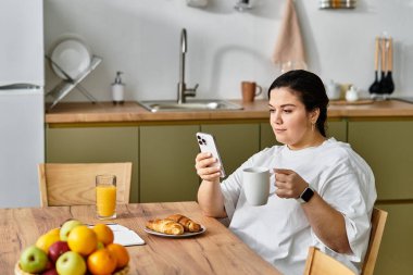 Parlak bir mutfakta akıllı telefonundaki mesajları dinlerken nefis bir kahvaltı yapıyor..