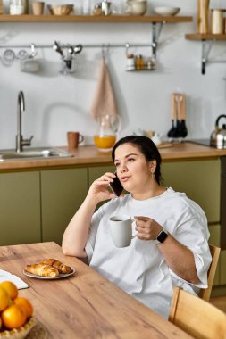 Genç ve iri yarı bir kadın, açık bir mutfakta kahvaltı ederken telefonla konuşmaktan zevk alıyor..