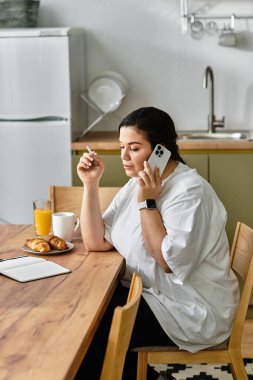 Young woman in a cozy kitchen talks on the phone while savoring breakfast and sipping juice. clipart