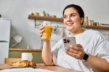 Neşeli genç bir kadın, güneşli bir mutfakta telefonunu kontrol ederken taze meyve suyu içiyor..