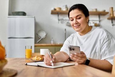 Genç bir kadın, ahşap bir masada, elinde telefonla defterine yazıyor..