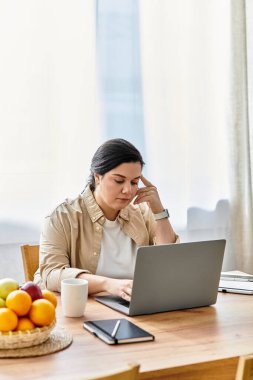 A young plus sized woman focuses on her laptop while surrounded by vibrant fruits in a cozy kitchen. clipart