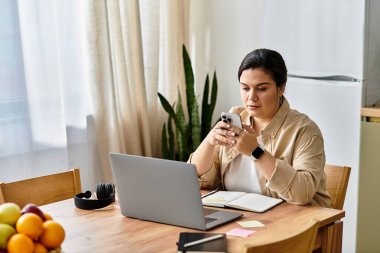 Genç ve iri yarı bir kadın ahşap bir masada oturuyor, dizüstü bilgisayarı ve telefonuyla özenle çalışıyor..