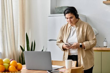 Genç ve iri yarı bir kadın ahşap bir masada duruyor. Telefonunu kullanıyor ve dizüstü bilgisayarı üzerinde çalışıyor..
