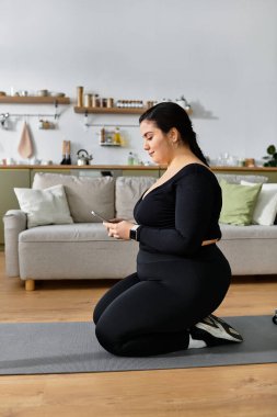 Büyük beden bir kadın, rahat bir oturma odasında, akıllı telefonuna odaklanmış bir şekilde paspasının üzerine diz çöker..