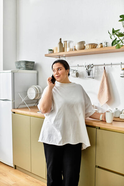 A young plus sized woman talks on the phone in a cozy, modern kitchen.