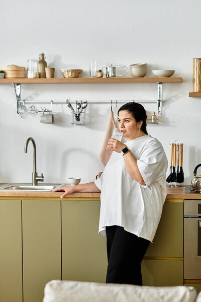 In a stylish kitchen, a plus sized woman stands sipping a drink, exuding confidence and tranquility.
