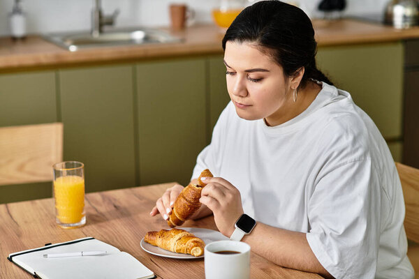 Молодая женщина плюс размер вкушает круассаны и апельсиновый сок, сидя за деревянным столом.
