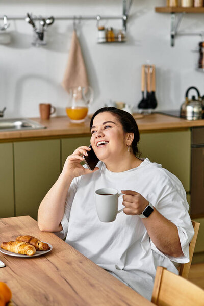 Молодая женщина плюс размер наслаждается кофе и выпечки за своим кухонным столом, болтая счастливо.
