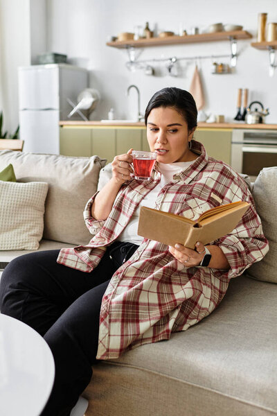 A young plus sized woman relaxes on a cozy couch, sipping tea while absorbed in a book.