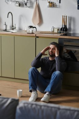 Yakışıklı bir adam mutfak zemininde oturuyor, düşüncelere dalmış, yanında bir fincan ile..