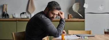 A thoughtful man sits at a wooden table, contemplating papers and looking at pills, lost in his thoughts. clipart