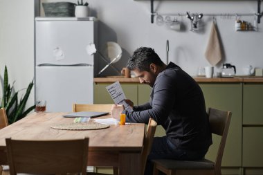 Yakışıklı bir adam ahşap bir masada oturuyor ve şık bir mutfakta önemli belgelerle uğraşıyor..
