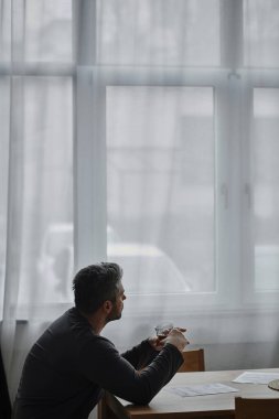 Düşünceli bir adam sıcak bir içeceği yudumlarken pencereden dışarı bakar..