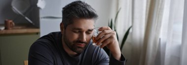 Yakışıklı bir adam düşünceli bir şekilde oturur, elinde bir bardak, evde dalgın dalgın dalgın oturur..