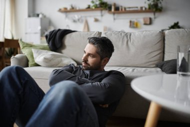 A thoughtful man sits on the floor of a stylish living room, gazing into the distance. clipart