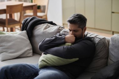 Bir adam kanepede dinleniyor, yastığını yakın tutarken huzurlu bir yerde düşüncelerde kayboluyor..