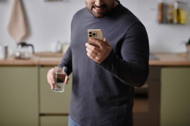 Yakışıklı bir adam elinde bir bardak su tutarken gülümser ve modern bir mutfakta telefonunu kontrol eder..