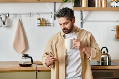 A handsome man sips coffee from a cup while looking at his phone in a modern kitchen setting. clipart