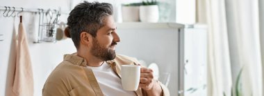 Yakışıklı bir adam ışıkla dolu modern bir mutfakta dinlenirken bir fincan sıcak kahvenin tadını çıkarıyor..