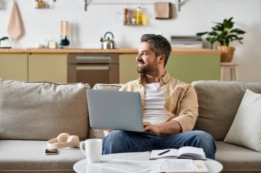 Yakışıklı bir adam rahat bir şekilde kanepede oturur, şık bir oturma odasında laptopuna odaklanır..