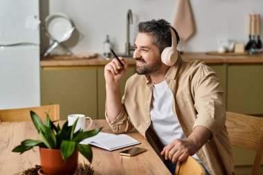 Handsome man smiles while listening to music and jotting ideas in his kitchen during the afternoon. clipart