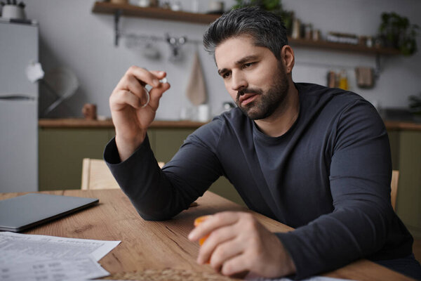 Красивый человек вдумчиво смотрит на таблетку, окруженную бумагами, в теплой кухне.