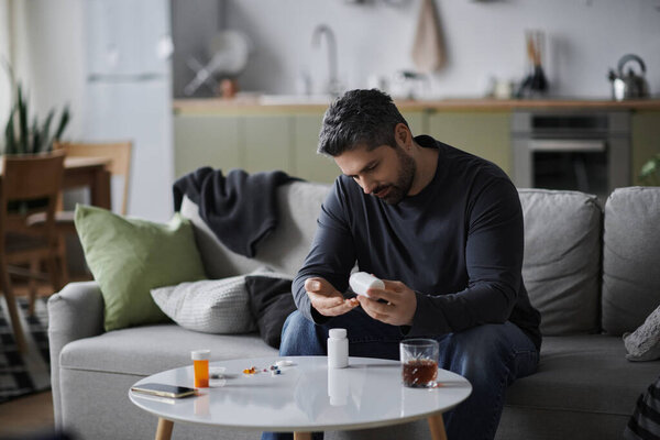 Красивый мужчина сидит на диване, размышляя о своем здоровье, рассматривая лекарства в уютной обстановке..