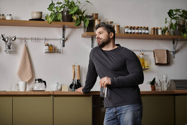 A handsome man leans against the counter in a modern kitchen, lost in thought with a glass.