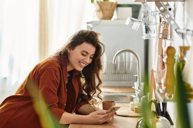 Bir kadın telefonunu kullanırken hafifçe gülümsüyor. Mutfak konforuyla çevrili. Umudu yansıtıyor..