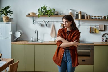 In a calm kitchen, a woman stands with crossed arms, showing her inner sadness. clipart