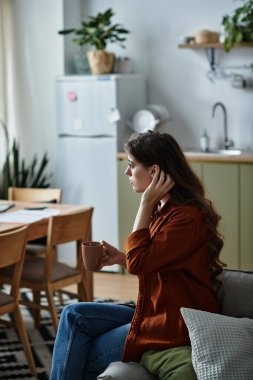 A woman sits pensively in a cozy kitchen, holding a warm cup, reflecting on her feelings of sadness. clipart