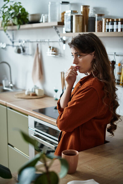 A woman stands in a serene kitchen, lost in thought as she reflects on her emotions.