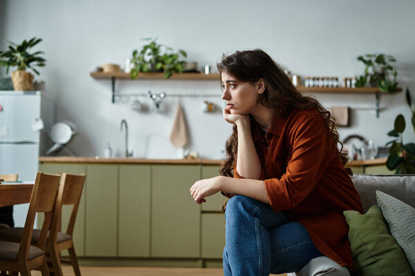 A woman sits on her couch, lost in thought, overwhelmed by sadness.