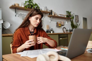 Mutfak masasında oturan bir kadın elinde bir kupayla belgelere bakarken dalgın dalgın bakıyor..
