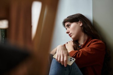 A young woman sits silently against a wall, lost in her thoughts and battling inner struggles. clipart