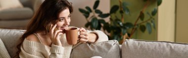 A woman clutches a steaming cup of tea, lost in thought within a serene living room. clipart