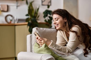 Genç bir kadın telefonuyla meşgul, rahat bir yerde otururken hafifçe gülümsüyor..