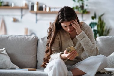 A woman sits on a couch, overwhelmed by emotions while looking at her phone in a cozy setting. clipart