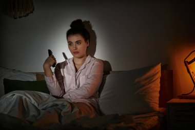 In a dimly lit bedroom, a woman sits on her bed, overwhelmed by feelings of depression and solitude. clipart