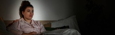 Genç bir kadın sessizce yatakta oturur, karanlık bir akşamda düşüncelerinde kaybolur, gölgelerle kaplanır..