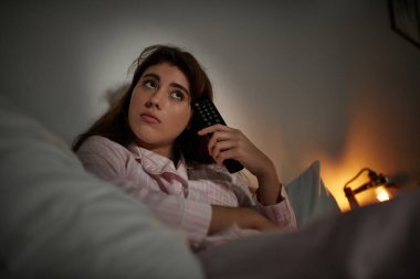 Uzaktan kumandalı bir bakışla düşüncelere dalmış bir kadın yatakta oturuyor, bakışlarında belli ki duygular var..