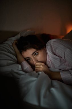 Bir kadın yatağına kıvrılmış, sorunlu düşüncelerinin ağırlığını yumuşak bir ışıltıyla sergiliyor..
