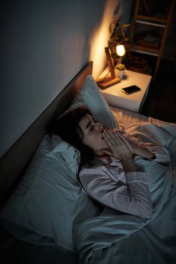 Bir kadın yatakta uzanmış, elleri dua ederken kenetlenmiş, gölgeler ve loş ışıklarla çevrili..