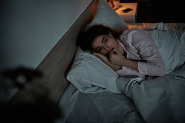 Bir kadın yan yatar, düşüncelere dalmış, yumuşak bir yatağın altında duygularının ağırlığını gösterir..